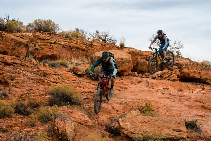Two riders on mountain bikes on red sandstone slickrock with desert shrubs