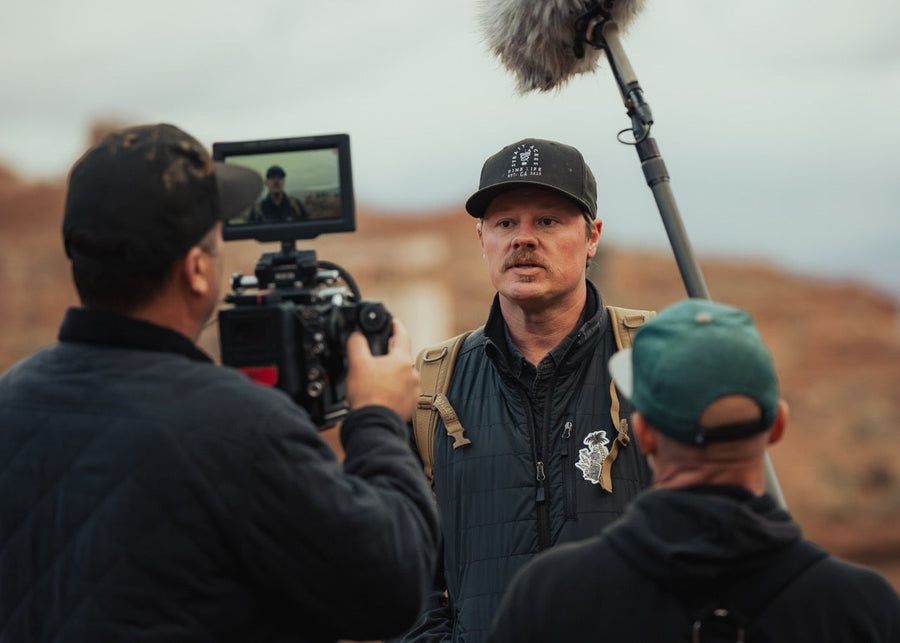 Man in a black cap being filmed on location, with a camera operator, boom mic and red rock behind