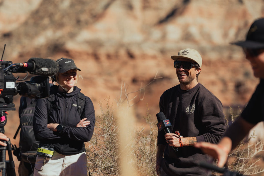Smiling man in a cap and sunglasses being interviewed with a microphone, camera crew beside him in red rock country