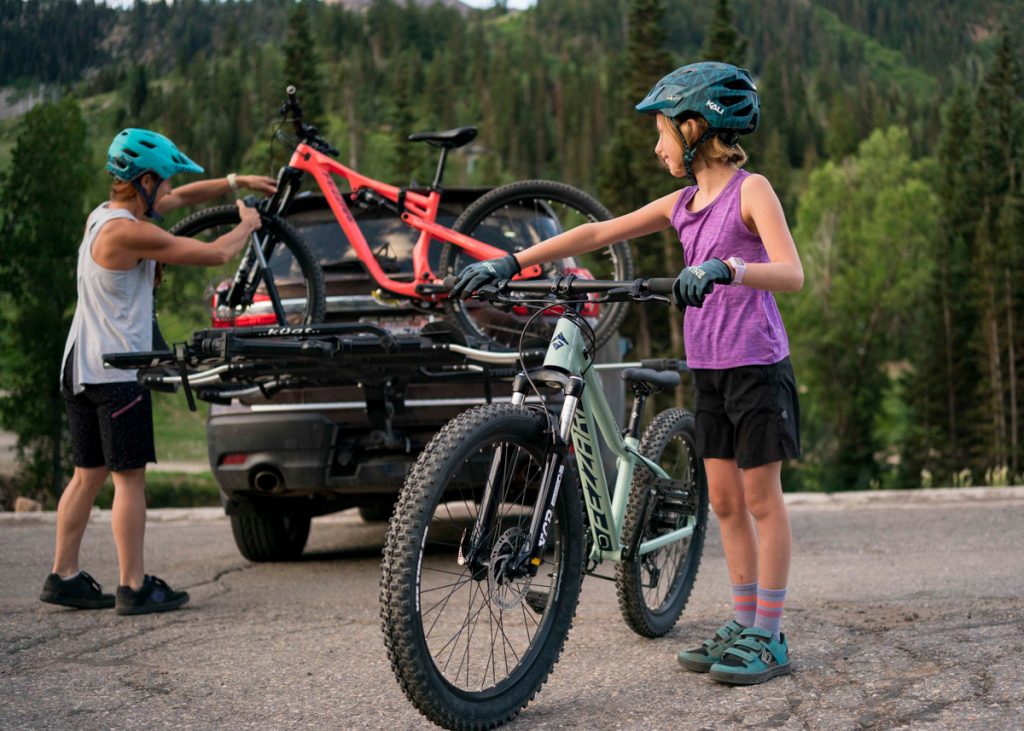 Two kids in teal helmets with kids' mountain bikes beside a car with one bike mounted on its rack