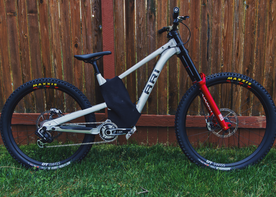 Silver Ari downhill electric bike with red fork and Maxxis tires leaning against a wooden fence