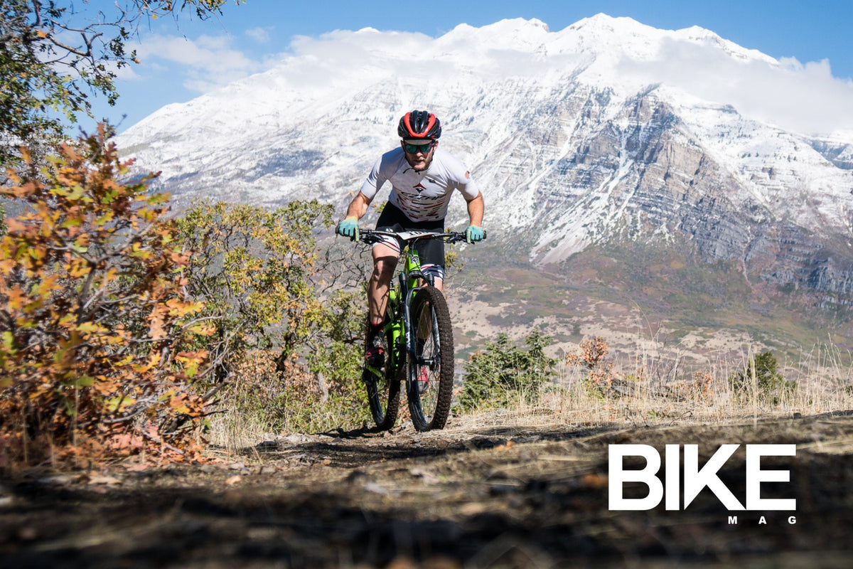 Rider on a green mountain bike on a trail below a snowy peak, with Bike Mag logo