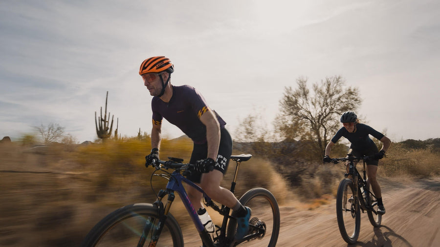 Two riders in dark jerseys on mountain bikes on a desert dirt road, one in an orange helmet