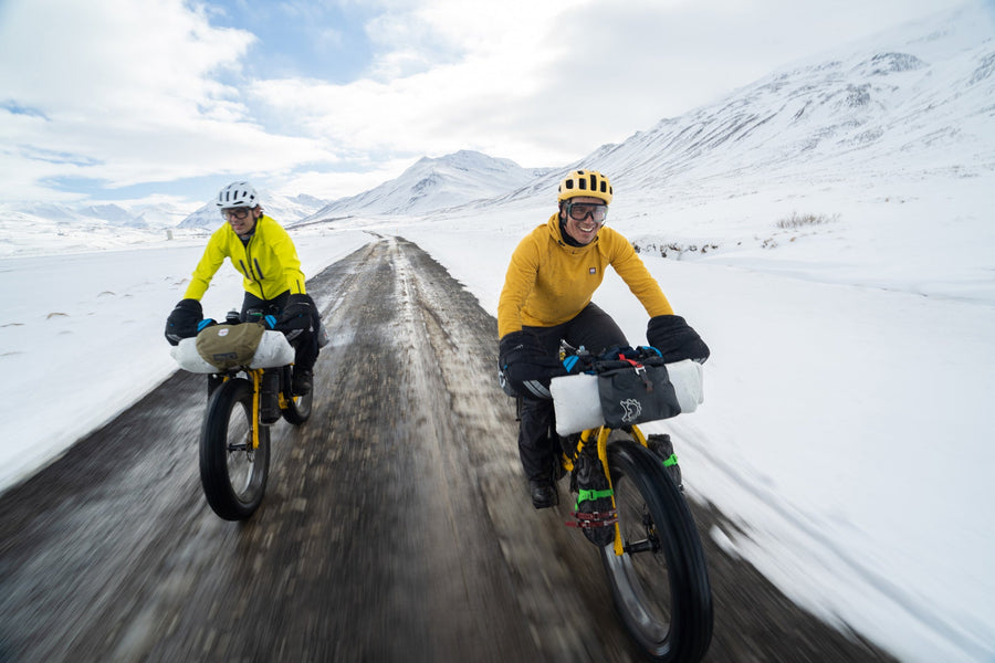 Two riders in yellow jackets on fat bikes with bikepacking bags on a muddy road through snowy Iceland