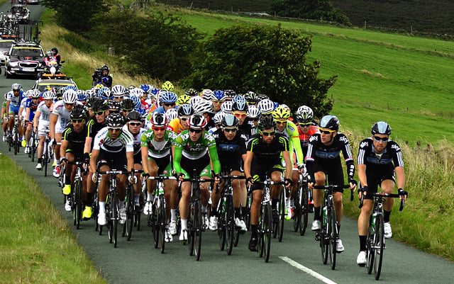 Peloton of road racers in team kits riding a country road with follow cars behind
