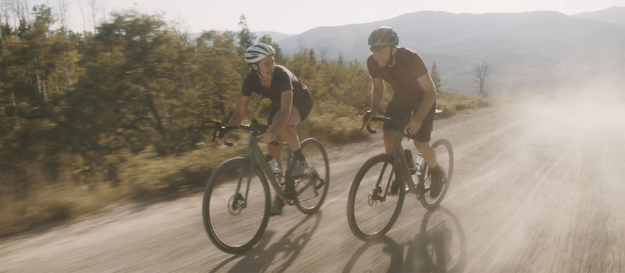 Two riders on gravel bikes on a dusty dirt road with trees and mountains behind