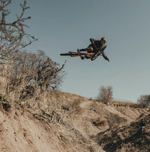 Rider whipping a mountain bike off a dirt jump against a gray sky on a dry hillside