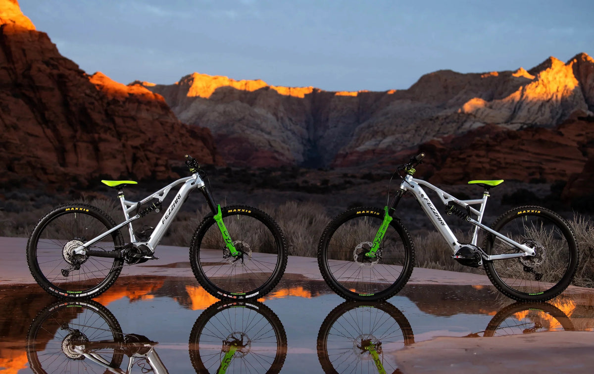 Two silver electric mountain bikes with green accents reflected in a pool in front of red rock cliffs