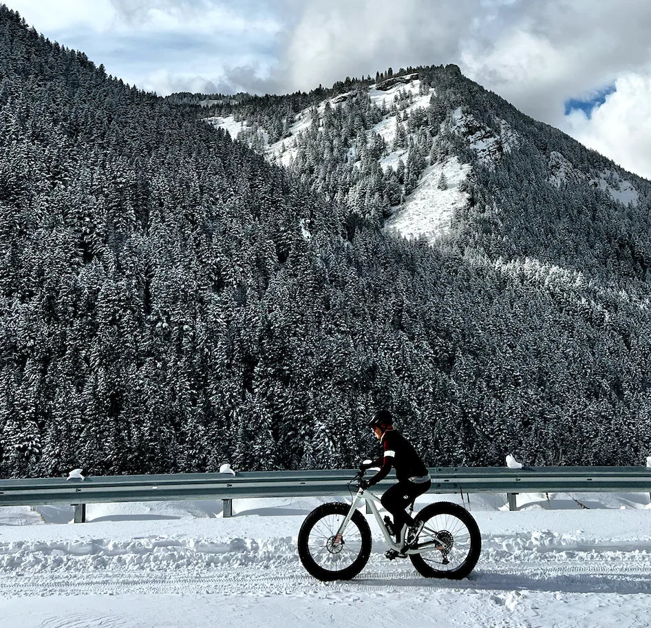 Rider on a fat bike on a snowy road below a snow-dusted forested mountain