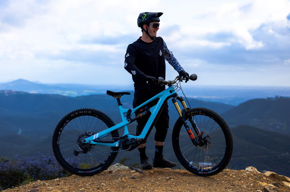 Nik Nestoroff standing with a light blue electric mountain bike on a hilltop overlooking a valley