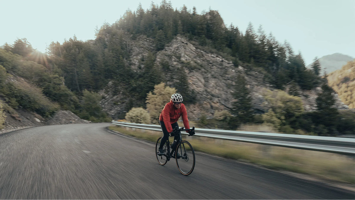 Rider in a red jacket on a road bike on a curving road beside a guardrail, rocky wooded hills behind