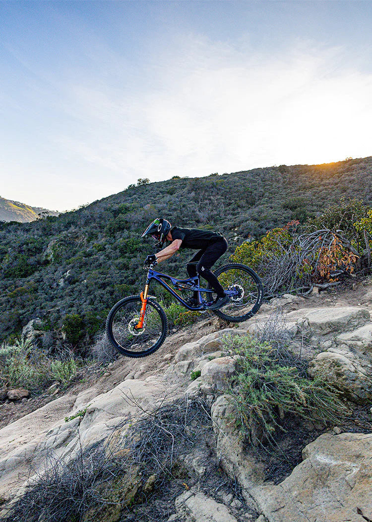 Rider on a blue full-suspension mountain bike with an orange fork descending a rocky hillside