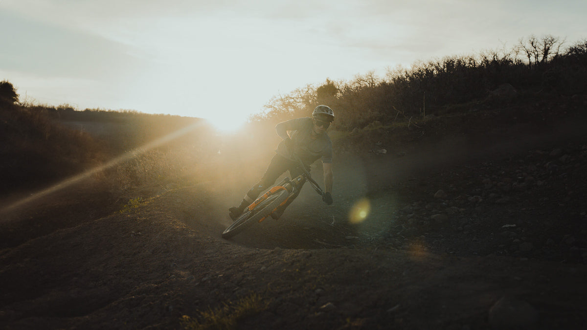 Silhouetted rider cornering a mountain bike on a dirt trail at sunset with sun flare