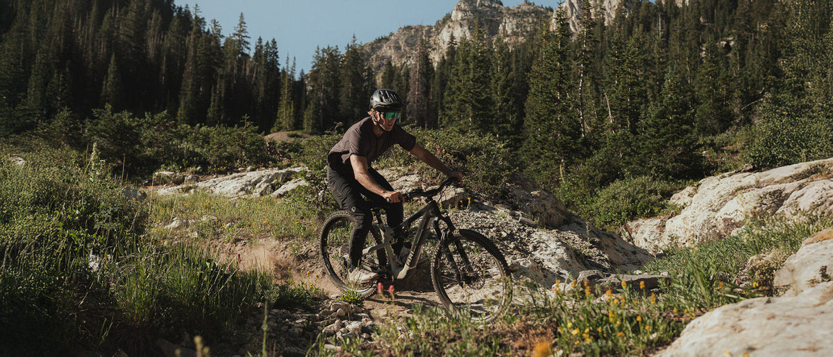 Rider on an electric mountain bike climbing a rocky trail with pine forest and cliffs behind