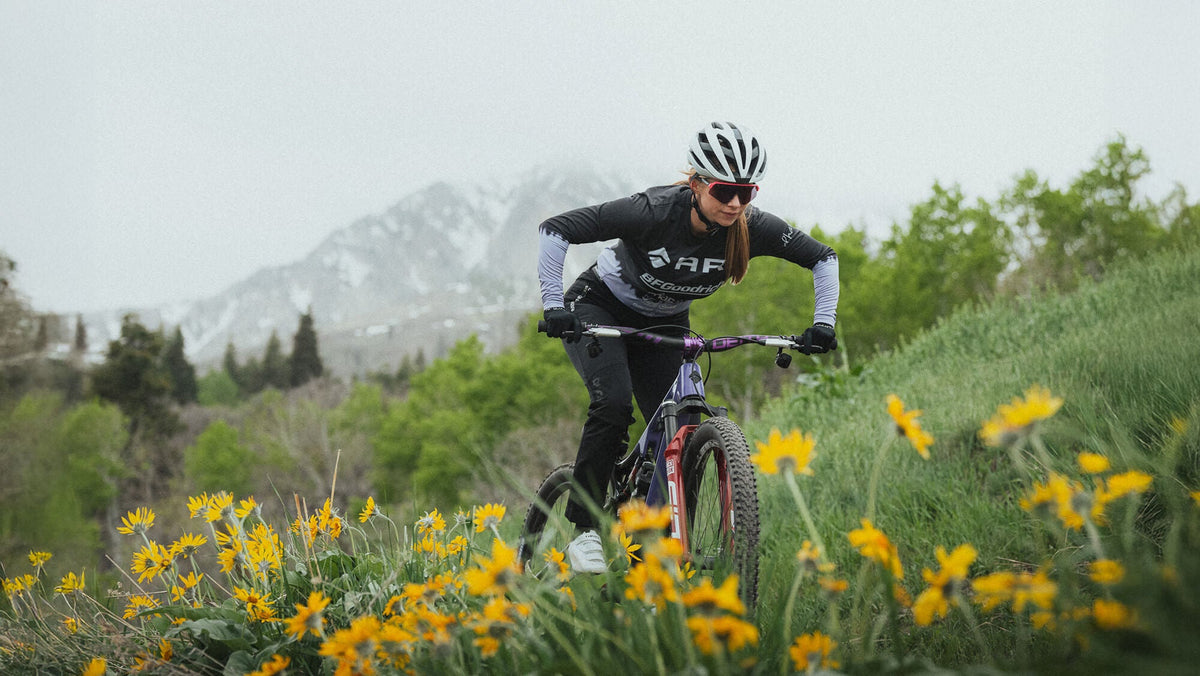 Rider in an Ari jersey on a mountain bike through yellow wildflowers with snowy peaks behind