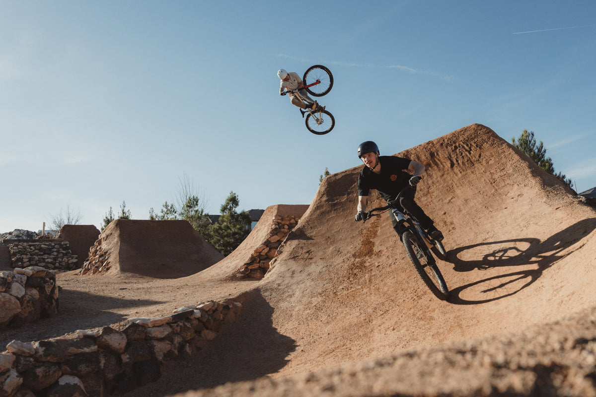 Rider carving a pump track berm on the 801 while another rider jumps overhead