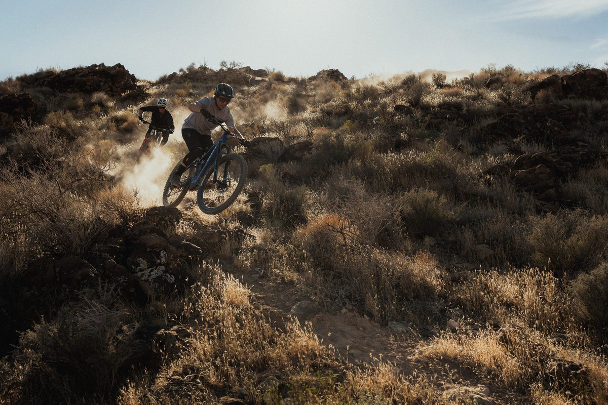 Rider kicking up dust descending a rocky desert trail with another rider behind