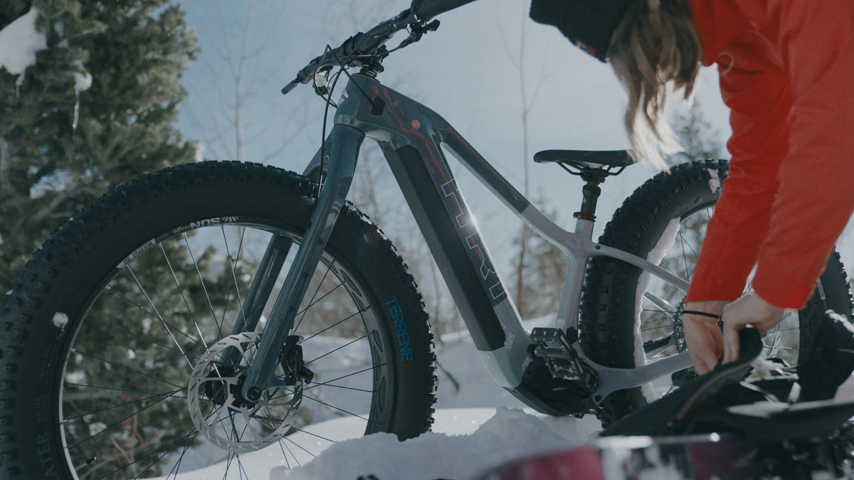 Rider in a red jacket adjusting a gray Explorer Peak fat e-bike in snow