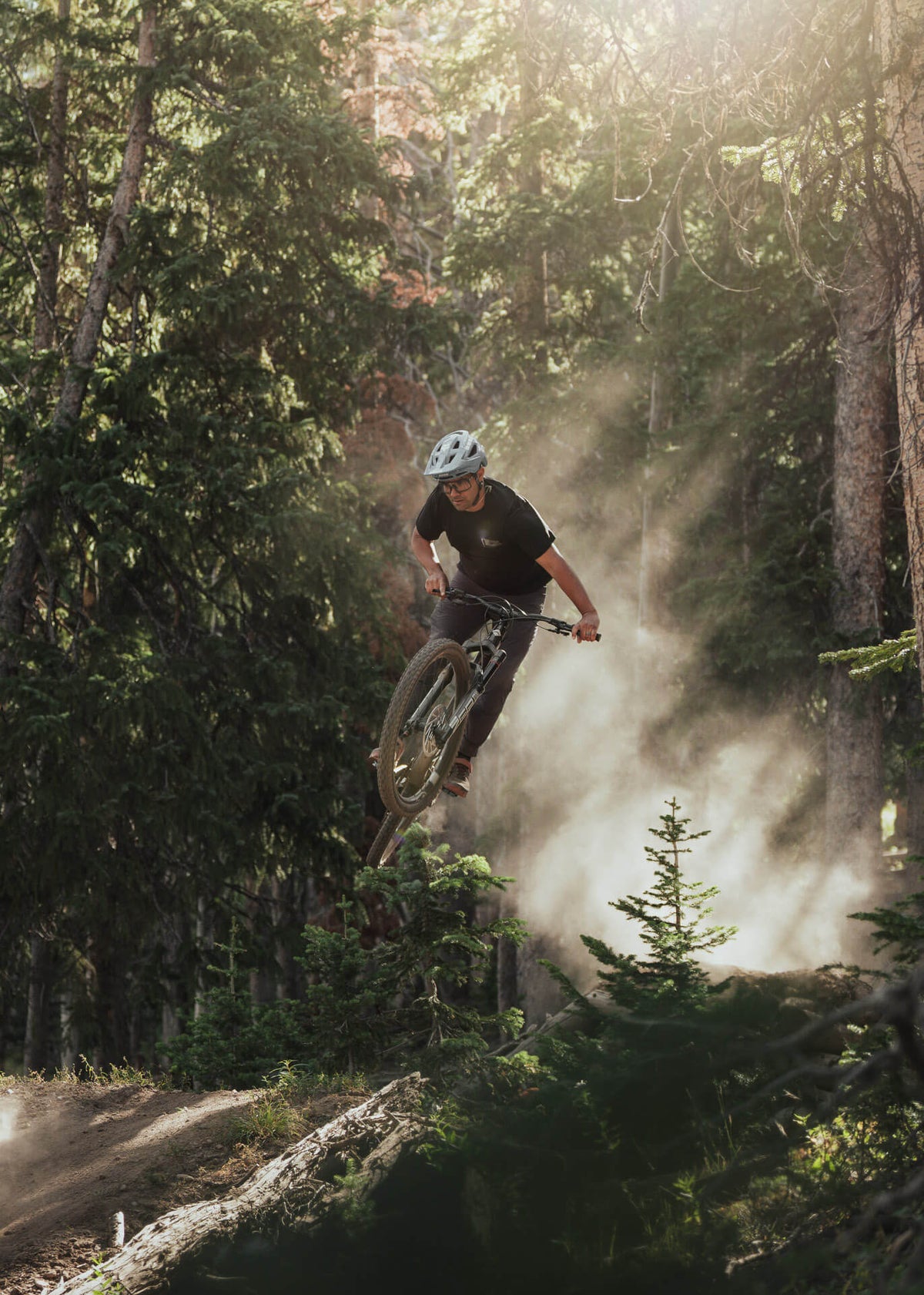 Rider jumping a mountain bike off a dirt lip in a sunlit forest with dust in the air