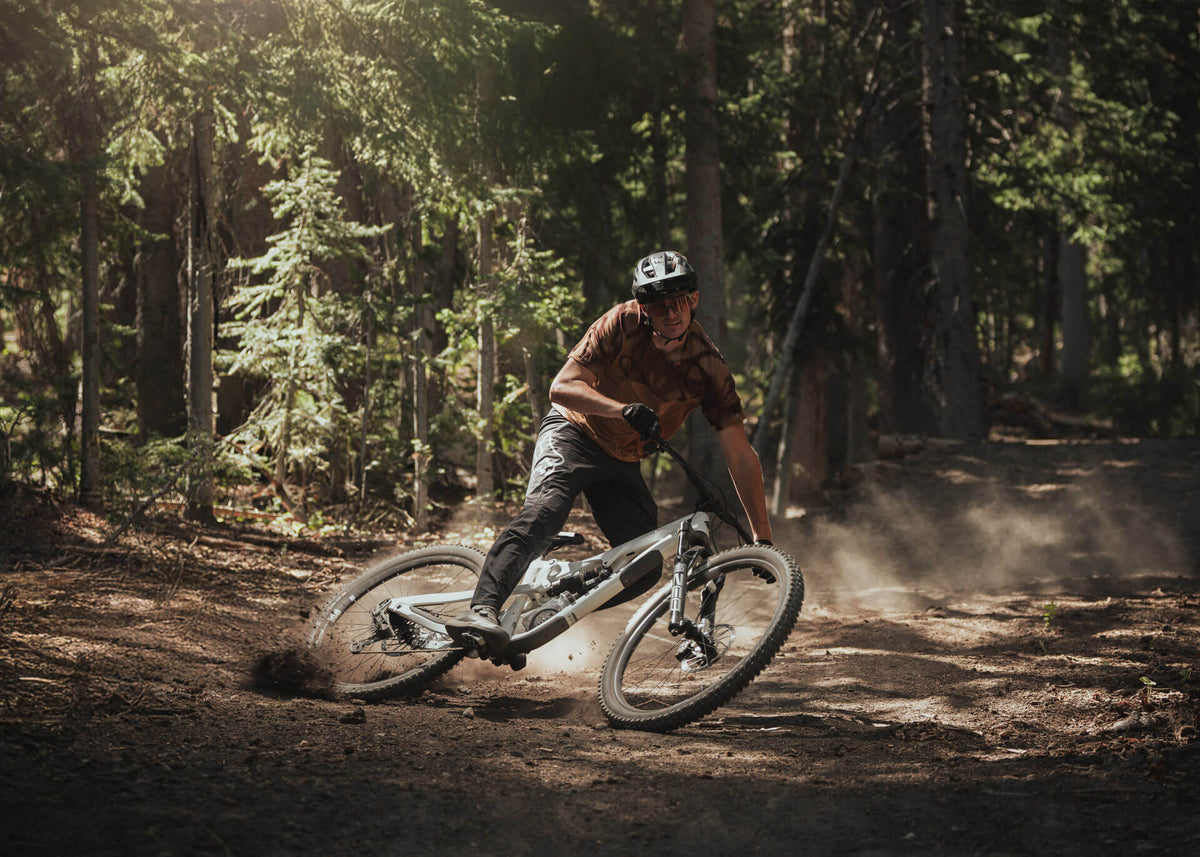 Rider sliding a corner on a dusty forest trail on a gray Ari full-suspension bike