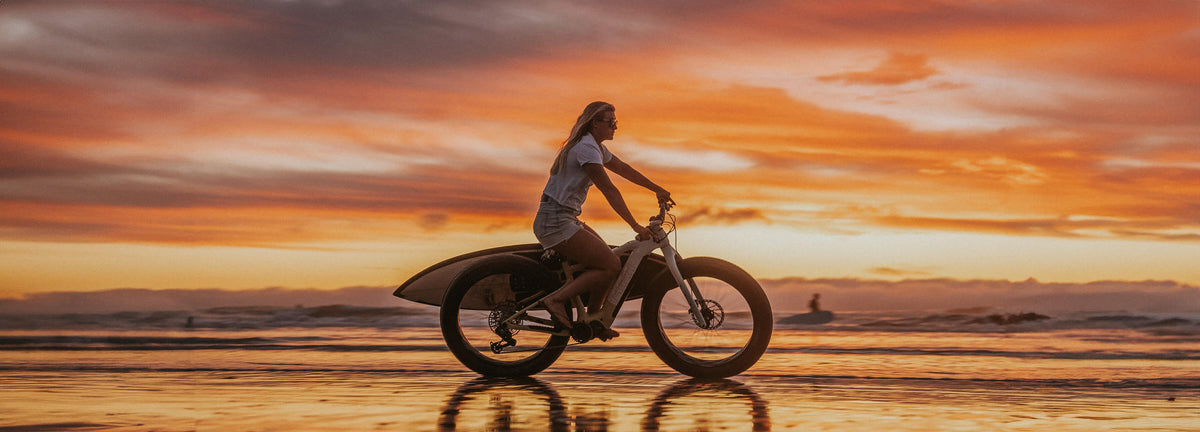 Woman riding a fat-tire e-bike on a beach at sunset with a surfboard on the rack