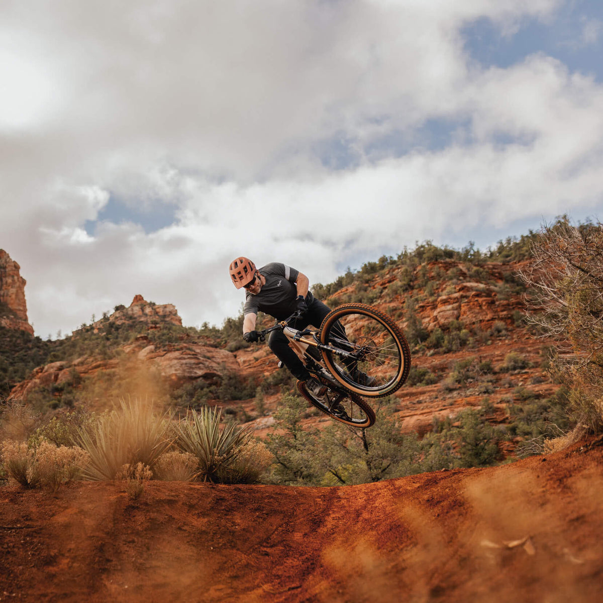 Rider jumping a mountain bike off red dirt with sandstone buttes behind