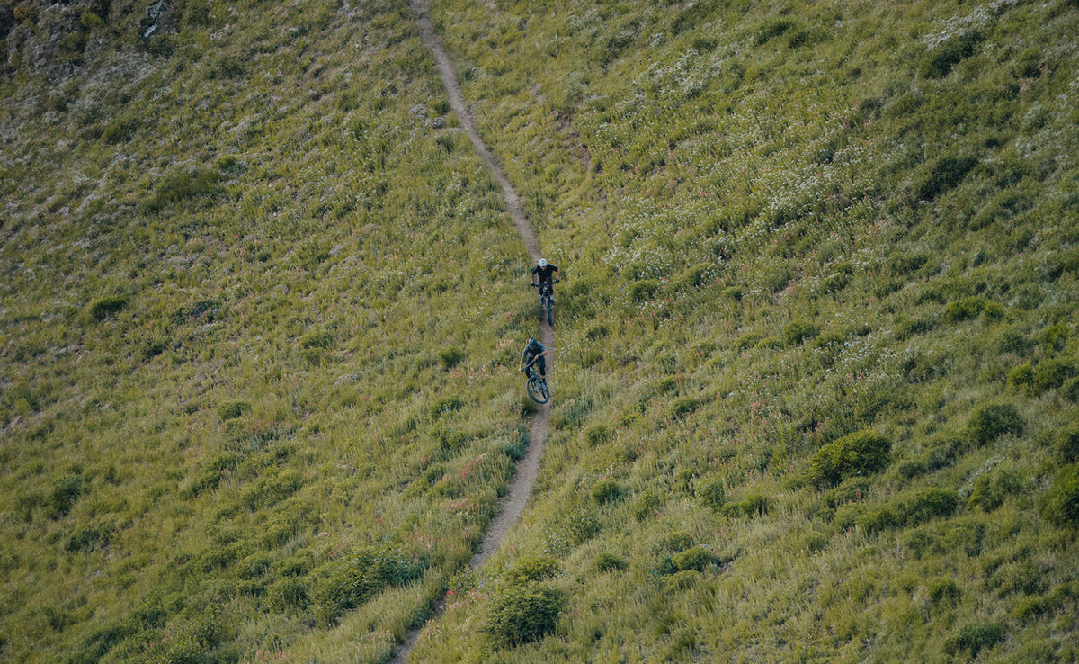 Aerial view of two riders descending a narrow singletrack across a grassy hillside