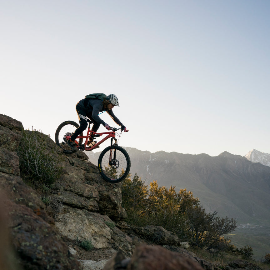 Rider descending a rocky ridge on a red full-suspension bike with mountains behind
