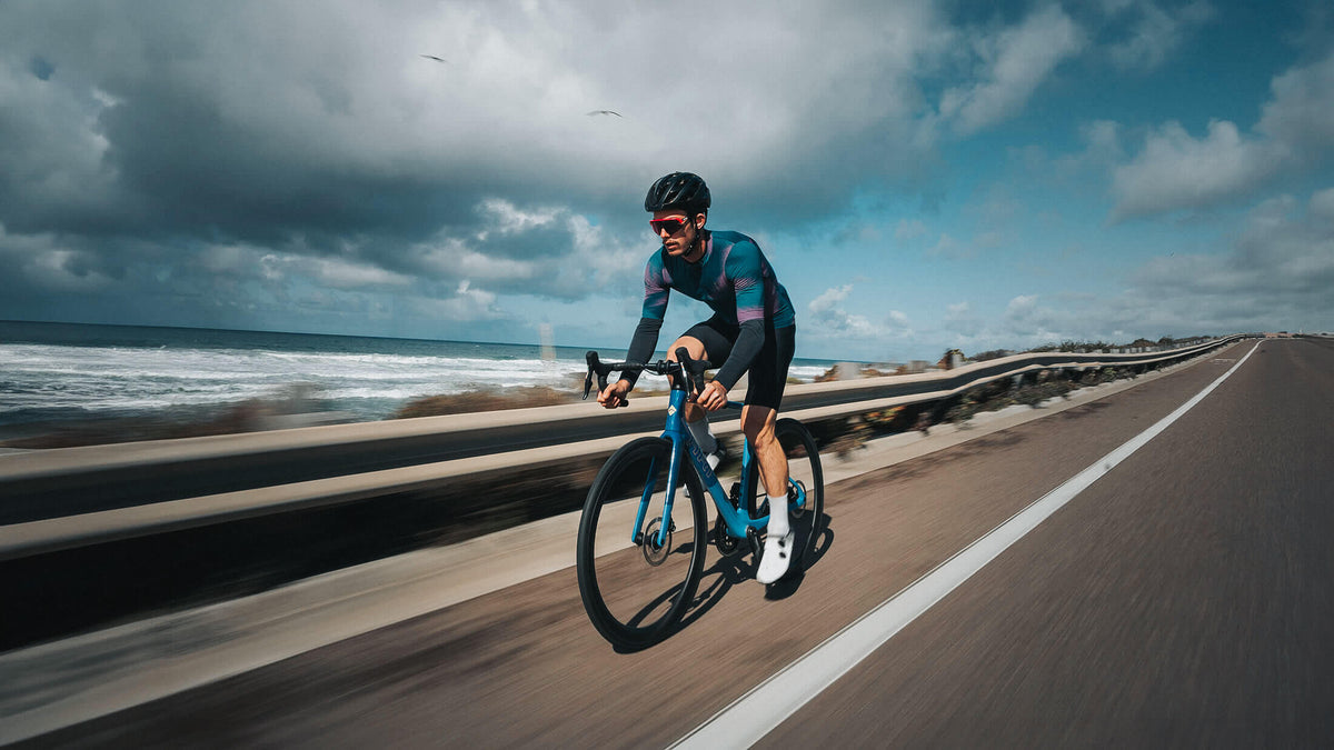Cyclist riding a blue road bike along a coastal road with waves beyond the guardrail