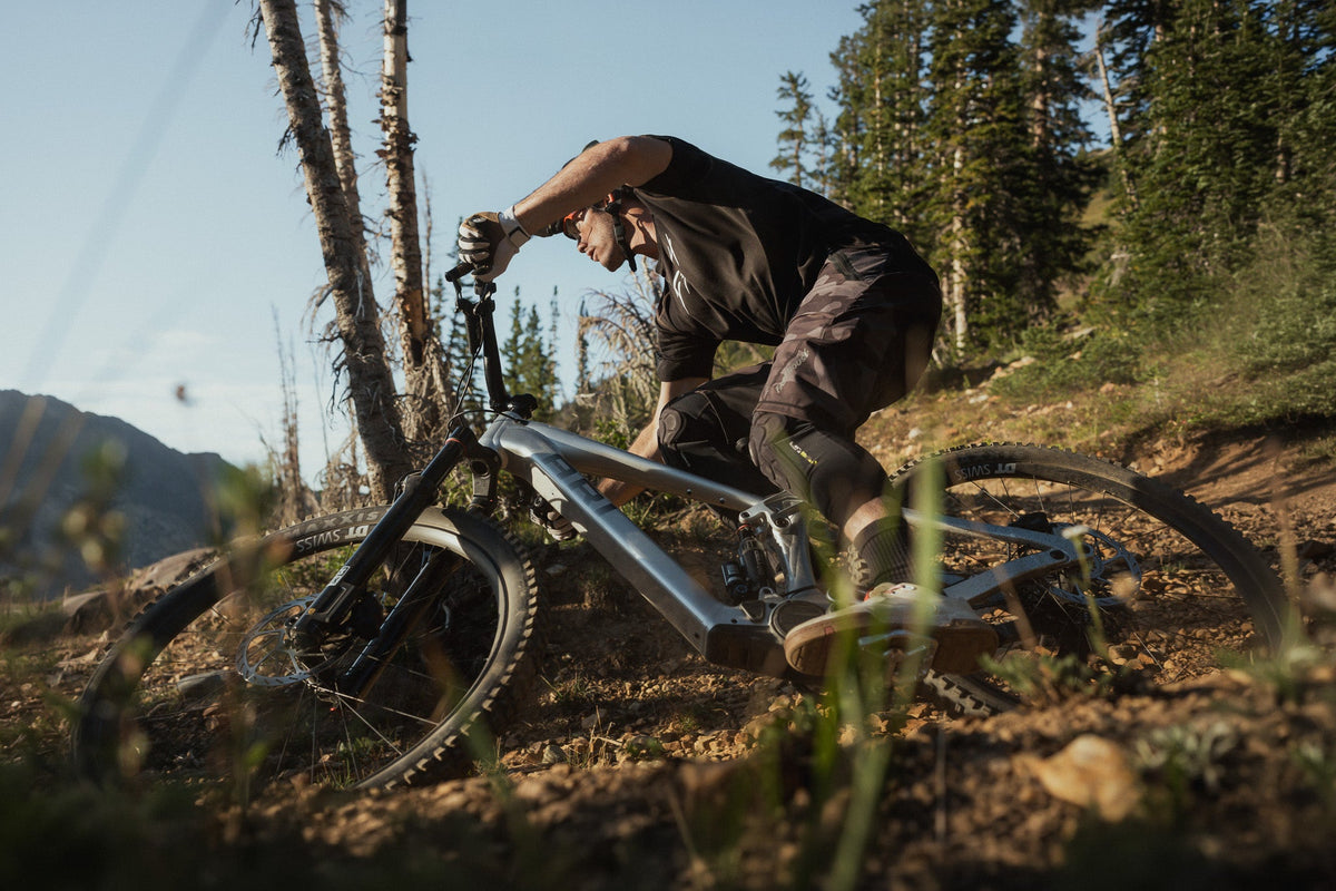 Rider tipping a Wire Peak e-bike into a loose dirt turn beside pine trees